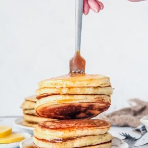 Three lemon pancakes are picked up with a fork whilst sitting in a stack on a white plate on a light grey surface.