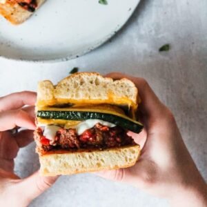 Meatball sandwich with slices of grilled zucchini and melted cheese are held over a grey background.