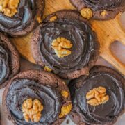 Afghan Biscuits rest on a wooden board on a light pink surface with swooshy chocolate frosting and walnuts in the center.