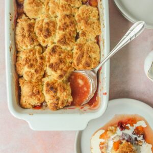 A serving spoon sits in the corner of a Plum and Apricot Cobbler in a white rectangle ceramic dish with a serving of cobbler with a drizzle of cream over the top on a light pink surface.