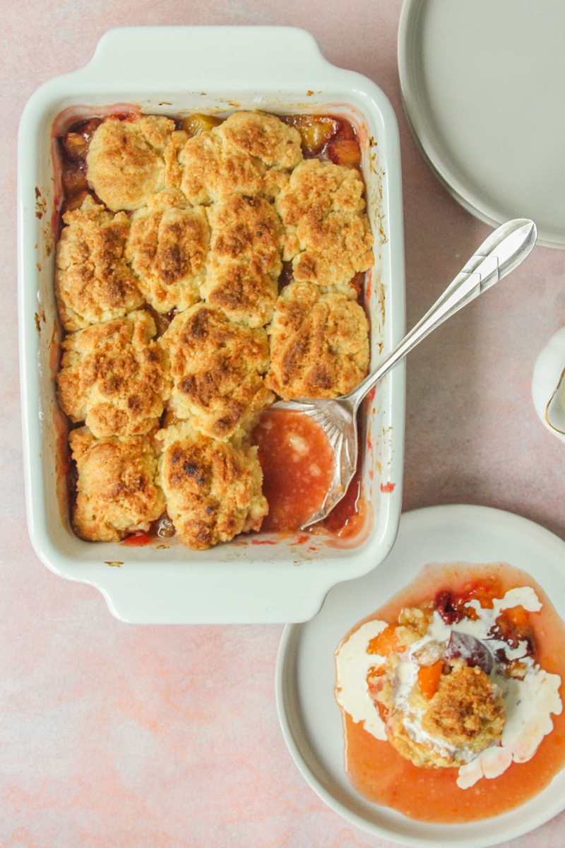 A serving spoon sits in the corner of a Plum and Apricot Cobbler in a white rectangle ceramic dish with a serving of cobbler with a drizzle of cream over the top on a light pink surface.