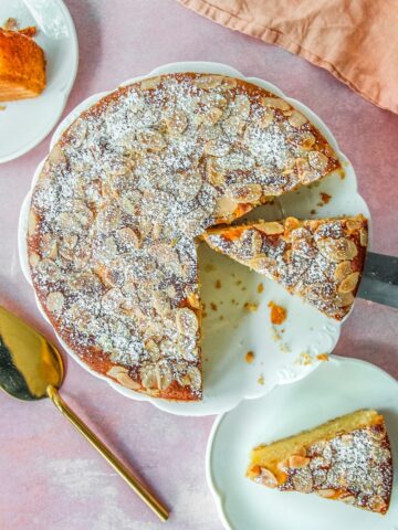 An Almond Croissant Cake sits on a fluted white ceramic plate with a couple of slices either side on a light pink surface.