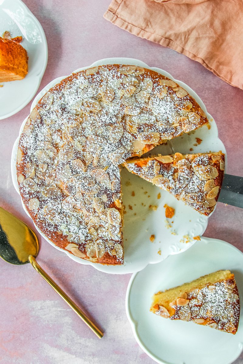 An Almond Croissant Cake sits on a fluted white ceramic plate with a couple of slices either side on a light pink surface.