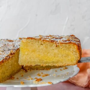An Almond Croissant Cake cake slice sits a cake server next to the remaining cake on a white ceramic fluted cake stand on a light pink surface.
