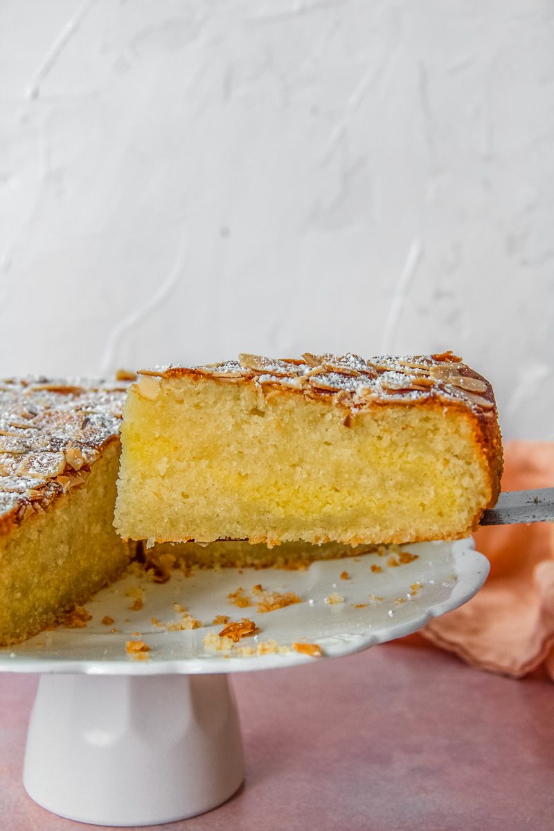 An Almond Croissant Cake cake slice sits a cake server next to the remaining cake on a white ceramic fluted cake stand on a light pink surface.