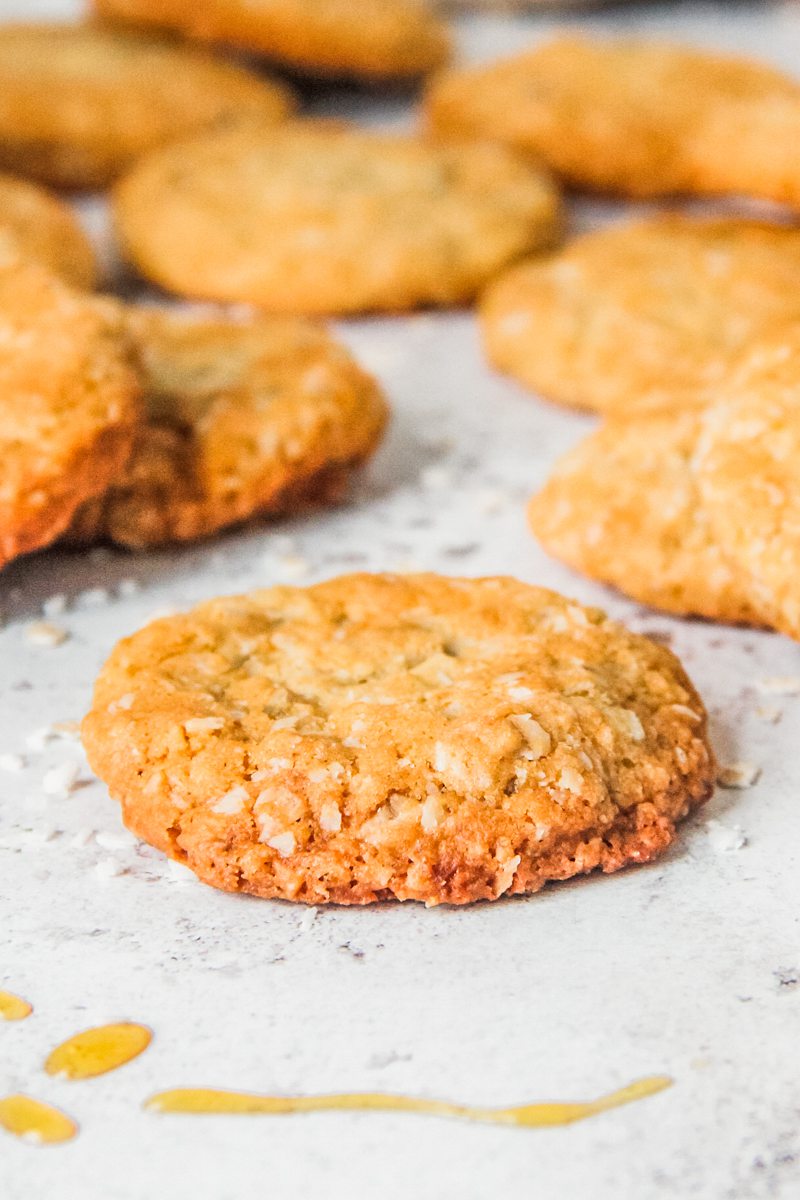 A close up of an Anzac biscuit laying on a light gray surface with other biscuits in the background.