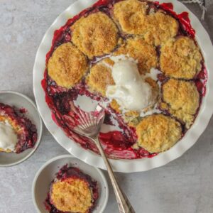 An apple berry cobbler topped with scoops of vanilla ice cream sits on a gray surface with two individual portions in gray ceramic bowls.