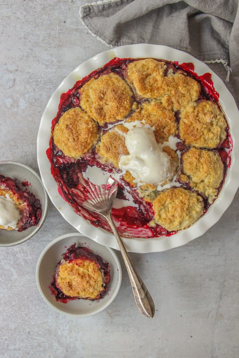 An apple berry cobbler topped with scoops of vanilla ice cream sits on a gray surface with two individual portions in gray ceramic bowls.