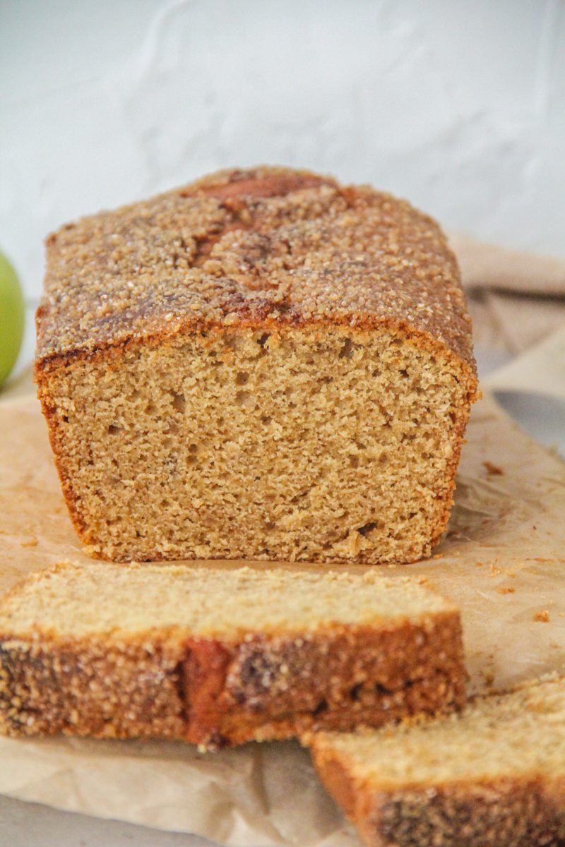 The light crumb of a Apple butter loaf cake is visible while sitting on parchment paper with a brown cloth behind.