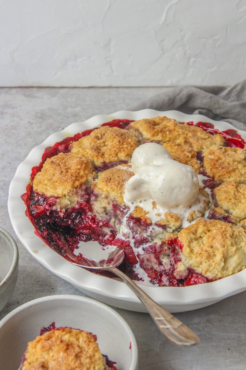 An apple berry cobbler topped with scoops of vanilla ice cream sits on a gray surface with individual portions in gray ceramic bowls.