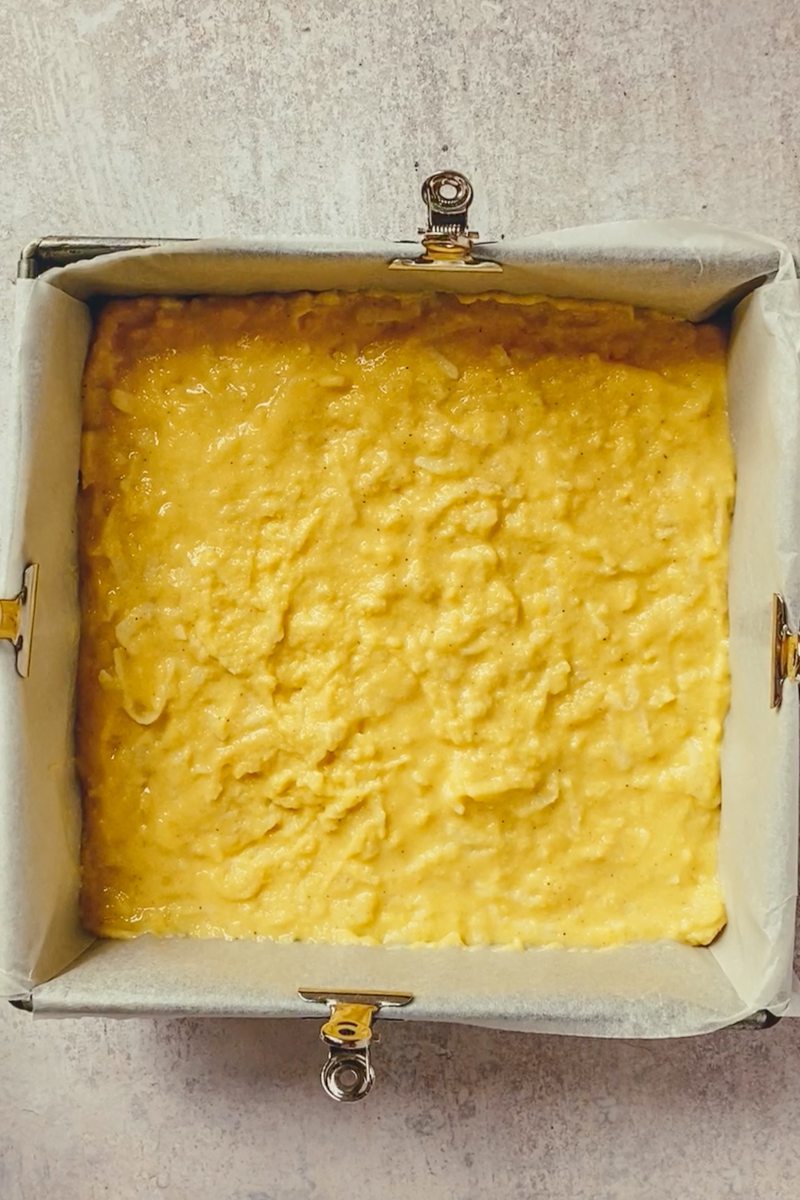 Unbaked apple cornbread batter sits in a lined square metal baking tin on a gray surface.