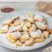 A side images of Austrian Kaiserschmarrn torn pancakes with jam on the side sits on a pale blue ceramic plate on a gray surface.