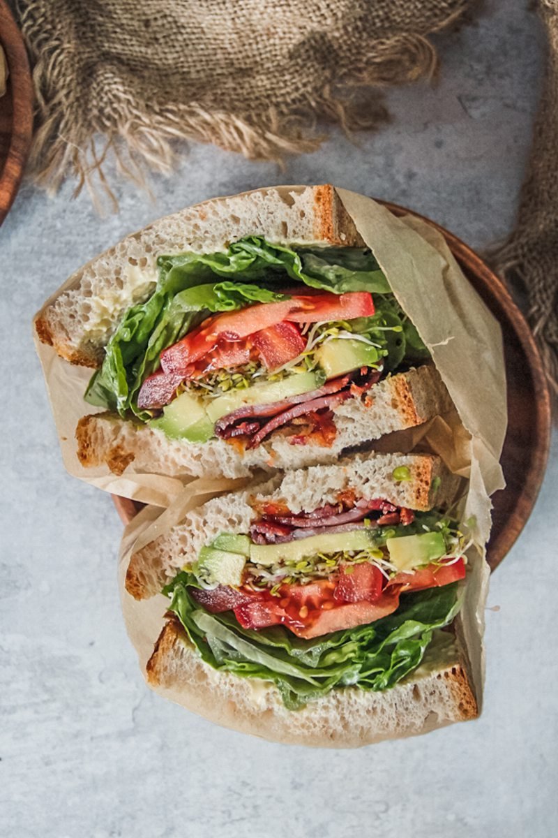 BLATS sandwich sits stacked on each other with the filling visible, on a wooden plate on a gray surface with a brown cloth beside.