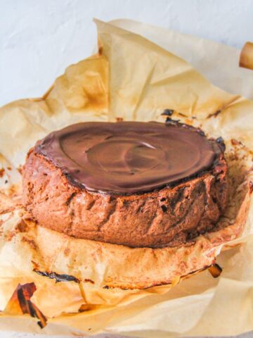 A chocolate Basque cheesecake sits on a cake stand in it's parchment paper wrapping on a light gray surface.