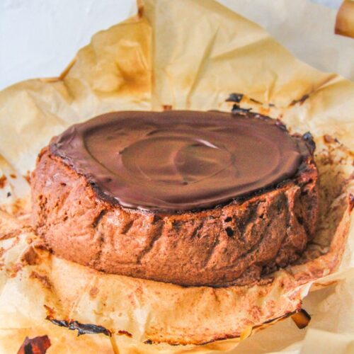 A chocolate Basque cheesecake sits on a cake stand in it's parchment paper wrapping on a light gray surface.