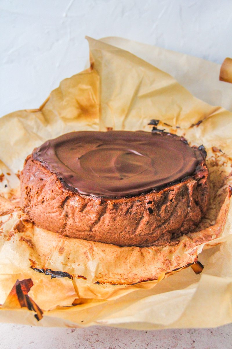 A chocolate Basque cheesecake sits on a cake stand in it's parchment paper wrapping on a light gray surface.
