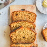 Banana Bread with Seed Brittle sits sliced on a wooden board with a slice and butter beside on a gray surface.