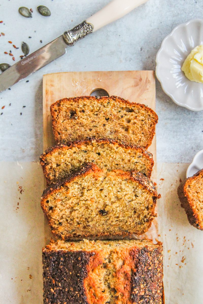 Banana Bread with Seed Brittle sits sliced on a wooden board with a slice and butter beside on a gray surface.