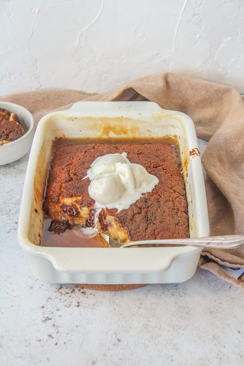 Banana Butterscotch pudding sits in a rectangle baking dish topped with scoops of vanilla ice cream on a light gray surface with a brown cloth behind.