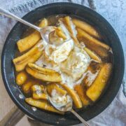 Banana's Foster sits in a saucepan with ice cream scoops and spoons resting in it on a gray surface.