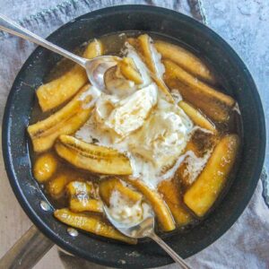 Banana's Foster sits in a saucepan with ice cream scoops and spoons resting in it on a gray surface.
