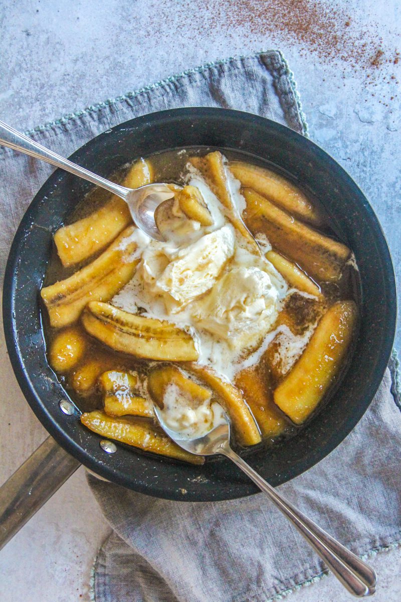 Banana's Foster sits in a saucepan with ice cream scoops and spoons resting in it on a gray surface.