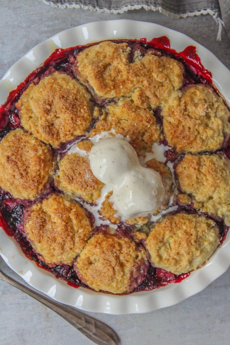 An apple berry cobbler topped with scoops of vanilla ice cream sits on a gray surface.