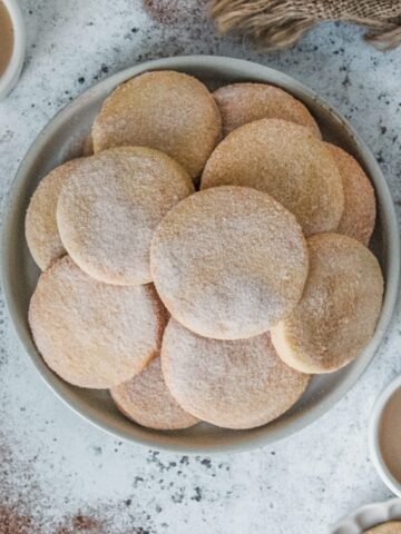 Biscochito cookies sit stacked on a rimmed ceramic gray plate on a gray surface.