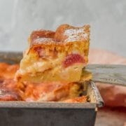 A slice of Estonian rhubarb cake sits on a cake server above the remaining cake in a square tin on a gray surface.