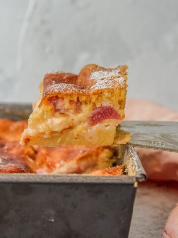 A slice of Estonian rhubarb cake sits on a cake server above the remaining cake in a square tin on a gray surface.