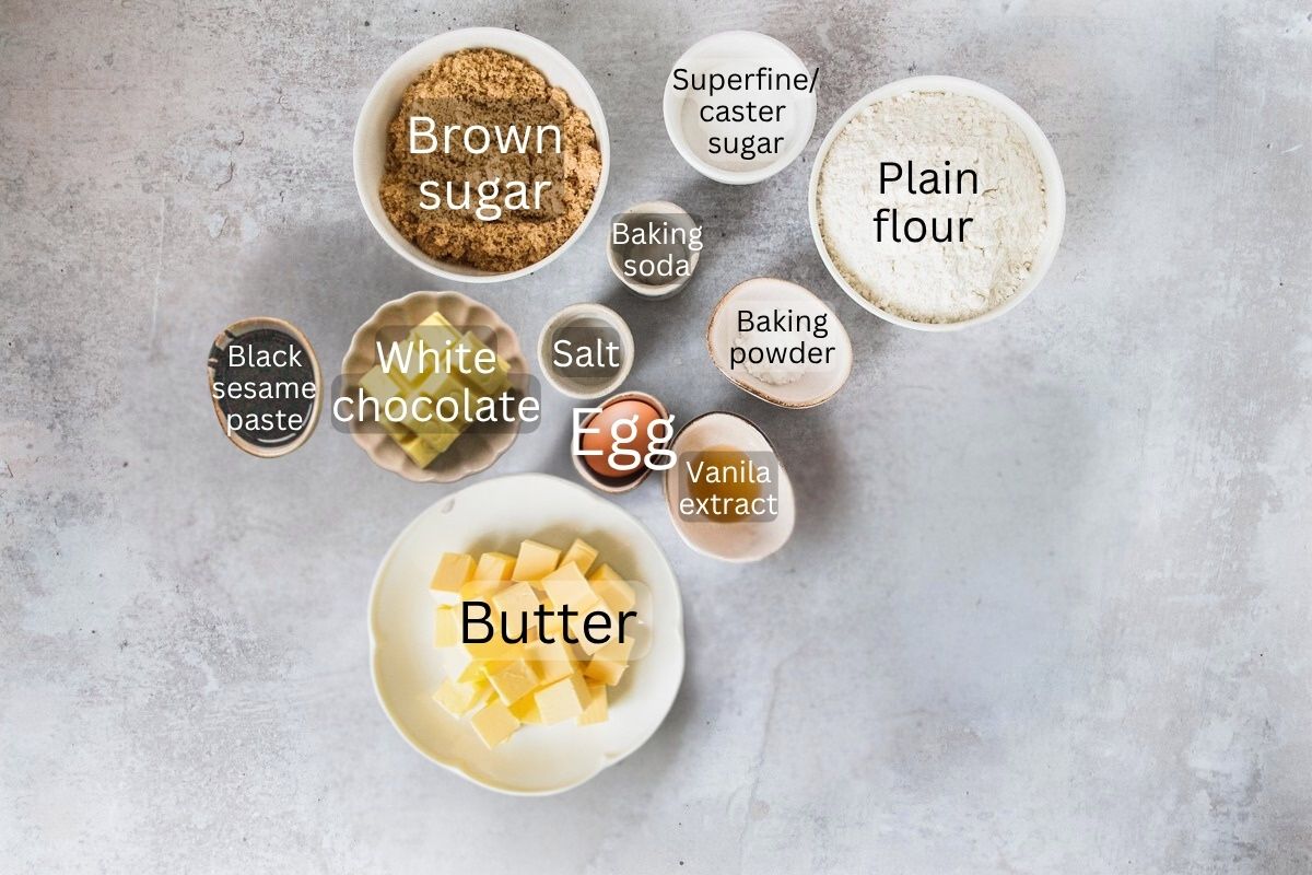 Ingredients for Black Sesame White Chocolate Cookies sit in a series of bowls and plates on a gray surface.