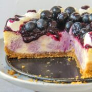 The interior of a swirled blueberry cheesecake with a jam and fresh berries on top and whipped cream on the edges on a cake stand on a gray surface.
