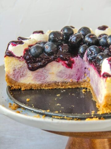 The interior of a swirled blueberry cheesecake with a jam and fresh berries on top and whipped cream on the edges on a cake stand on a gray surface.