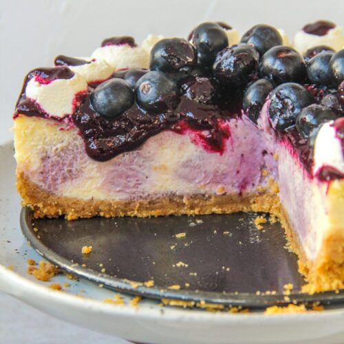 The interior of a swirled blueberry cheesecake with a jam and fresh berries on top and whipped cream on the edges on a cake stand on a gray surface.