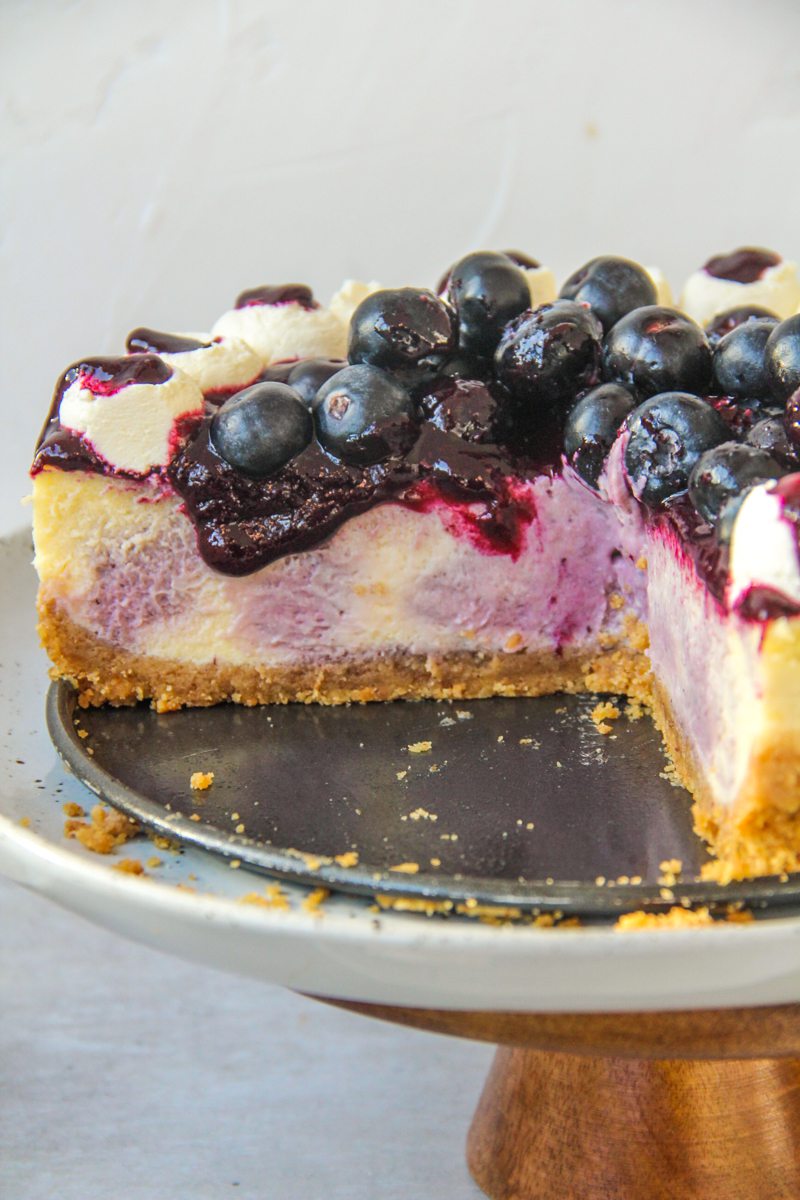 The interior of a swirled blueberry cheesecake with a jam and fresh berries on top and whipped cream on the edges on a cake stand on a gray surface.