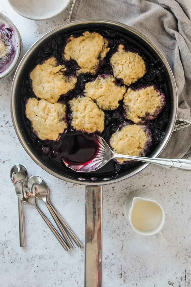 Blueberry Grunt or Canadian Blueberry Cobbler sits on a light grey background, served up in a stainless steel pan with a serving spoon