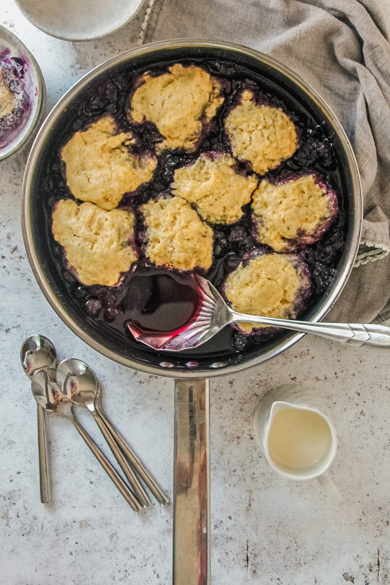 Canadian Blueberry grunt or Blueberry cobbler sits in a stainless steel saucepan on a light gray surface with serving spoons and a small jug of cream beside with a serving spoon stuck into the juice with an individual serving beside.