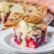 A slice of peach and blueberry pie sits on a stack of individual plates with a scoop of melting ice cream on top on a light gray surface.