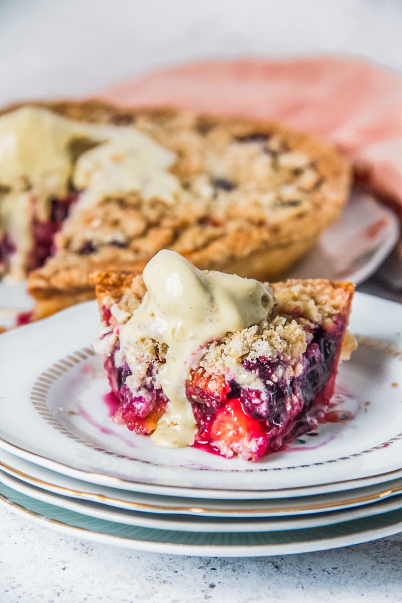 A slice of peach and blueberry pie sits on a stack of individual plates with a scoop of melting ice cream on top on a light gray surface.