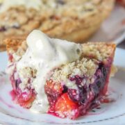 A slice of peach and blueberry pie sits on a stack of individual plates with a scoop of melting ice cream on top on a light gray surface.