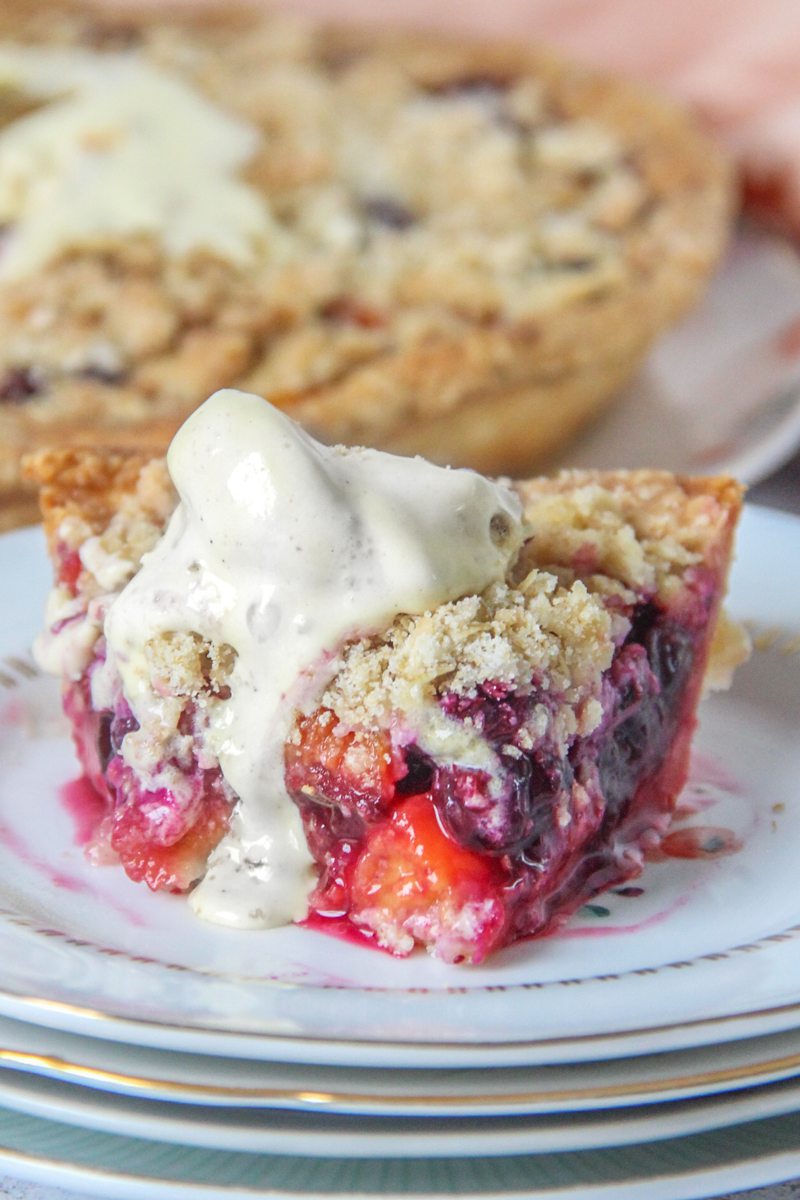A slice of peach and blueberry pie sits on a stack of individual plates with a scoop of melting ice cream on top on a light gray surface.