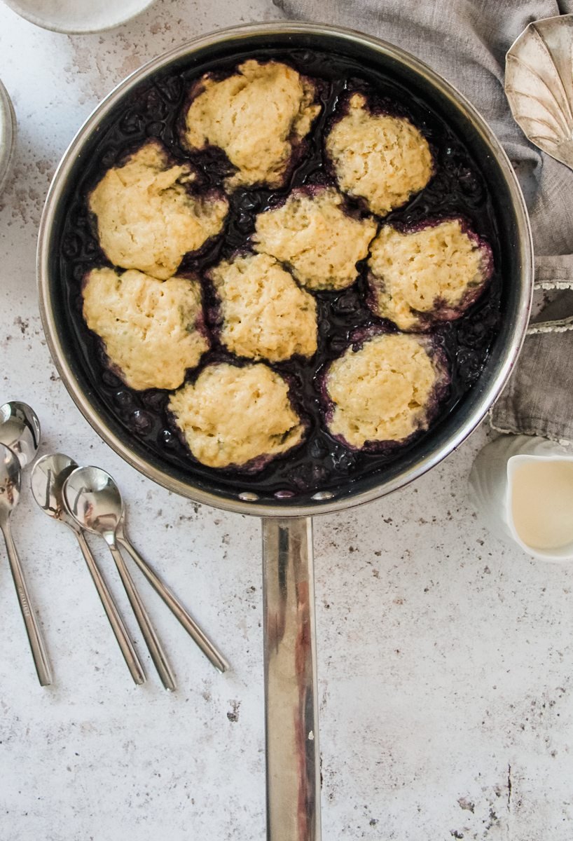 Blueberry Grunt or Canadian Blueberry Cobbler sits on a light grey background, served up in a stainless steel pan.