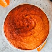 A whole orange cake sits on a fluted white ceramic plate on a light gray surface.
