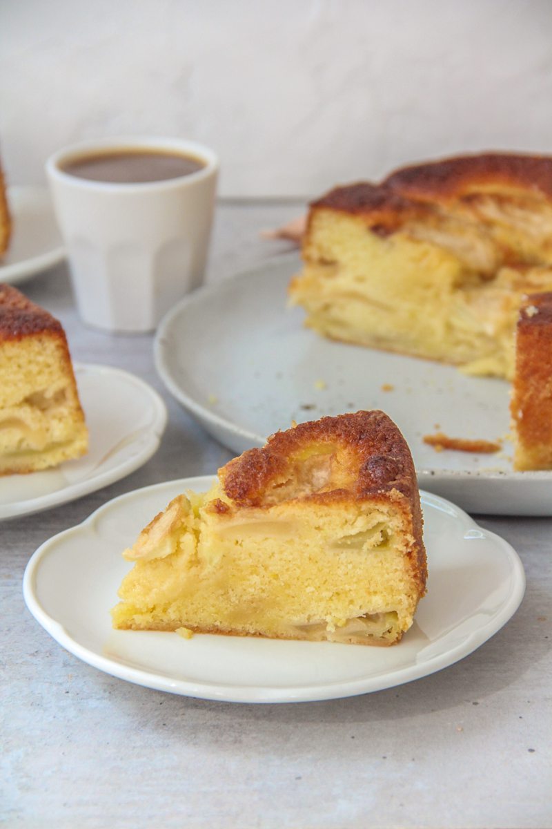 A slice of Dorset Apple Cake sits on a white ceramic plate on a grey surface with the remaining cake beside.