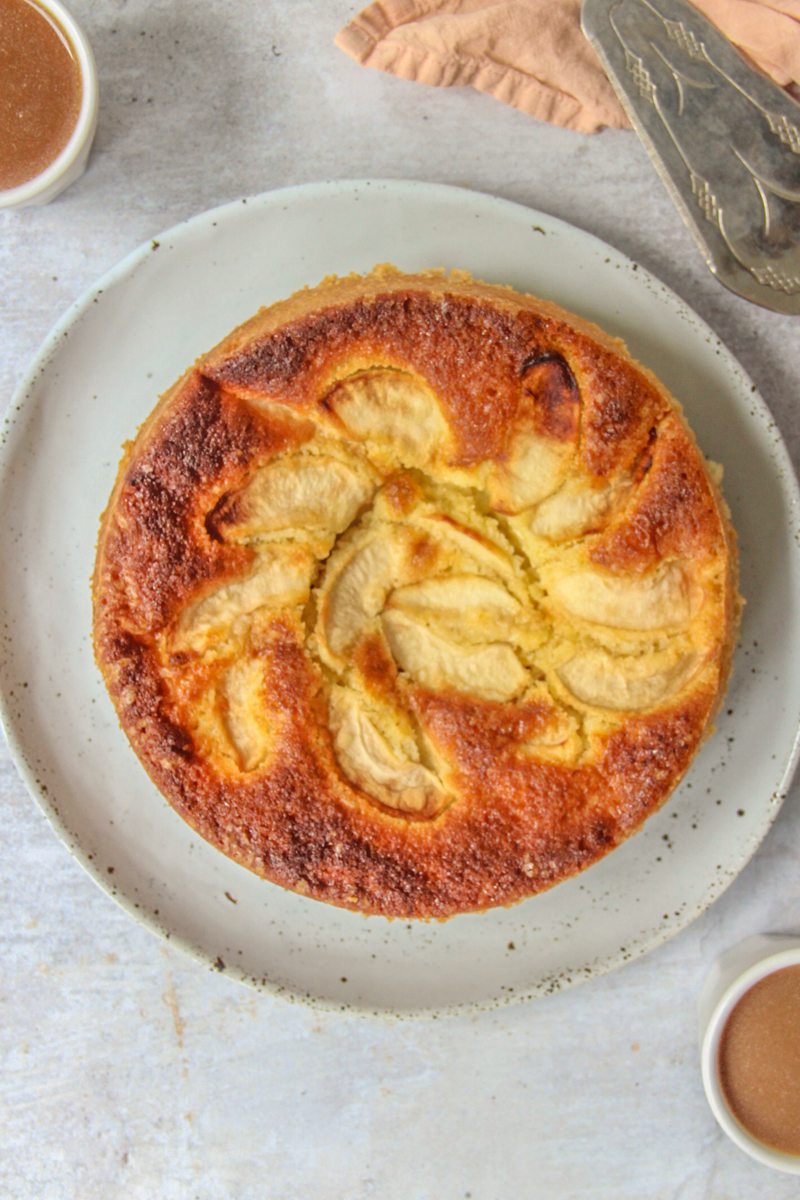 British Dorset Apple Cake sits on a light blue ceramic plate on a grey surface with two small cups of coffee beside.