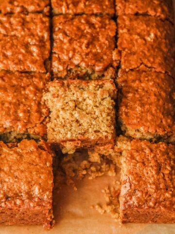 A slice of Ginger Parkin sits leaning against the remaining cake slices on a parchment lined surface.