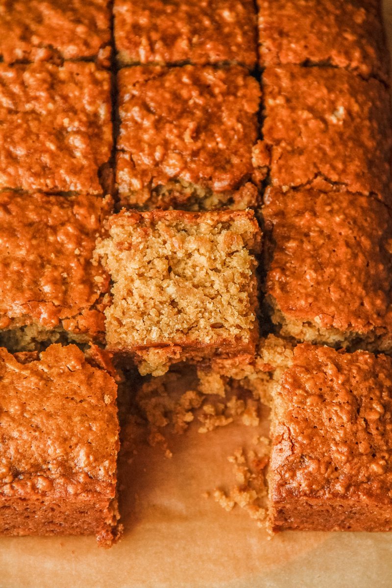 A slice of Ginger Parkin sits leaning against the remaining cake slices on a parchment lined surface.