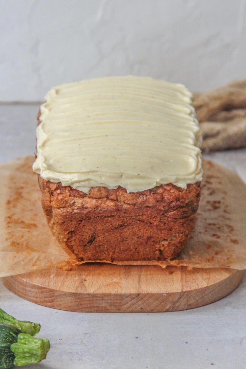 A zucchini loaf cake with brown butter cream cheese frosting sits on parchment paper on a wooden board on a gray surface.