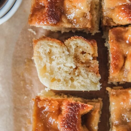 A slice of Danish Coffee Cake sits on it's side with the fluffy yeast interior visible surrounded by other pieces sitting upright and the poked holes and caramelized topping visible on a brown parchment paper surface.