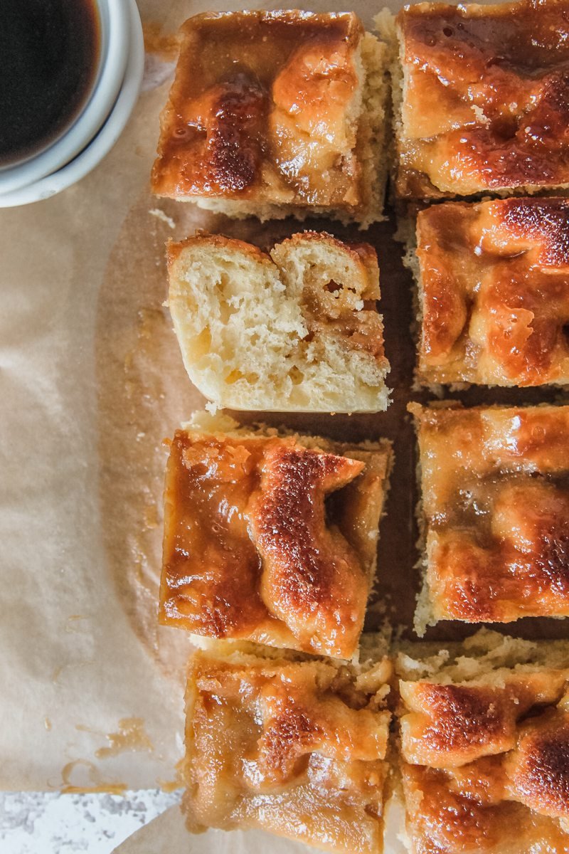 A slice of Brunsviger - Danish Coffee Cake sits on it's side with the fluffy yeast interior visible surrounded by other pieces sitting upright and the poked holes in the yeasted cake and caramelized topping visible on a brown parchment paper surface.